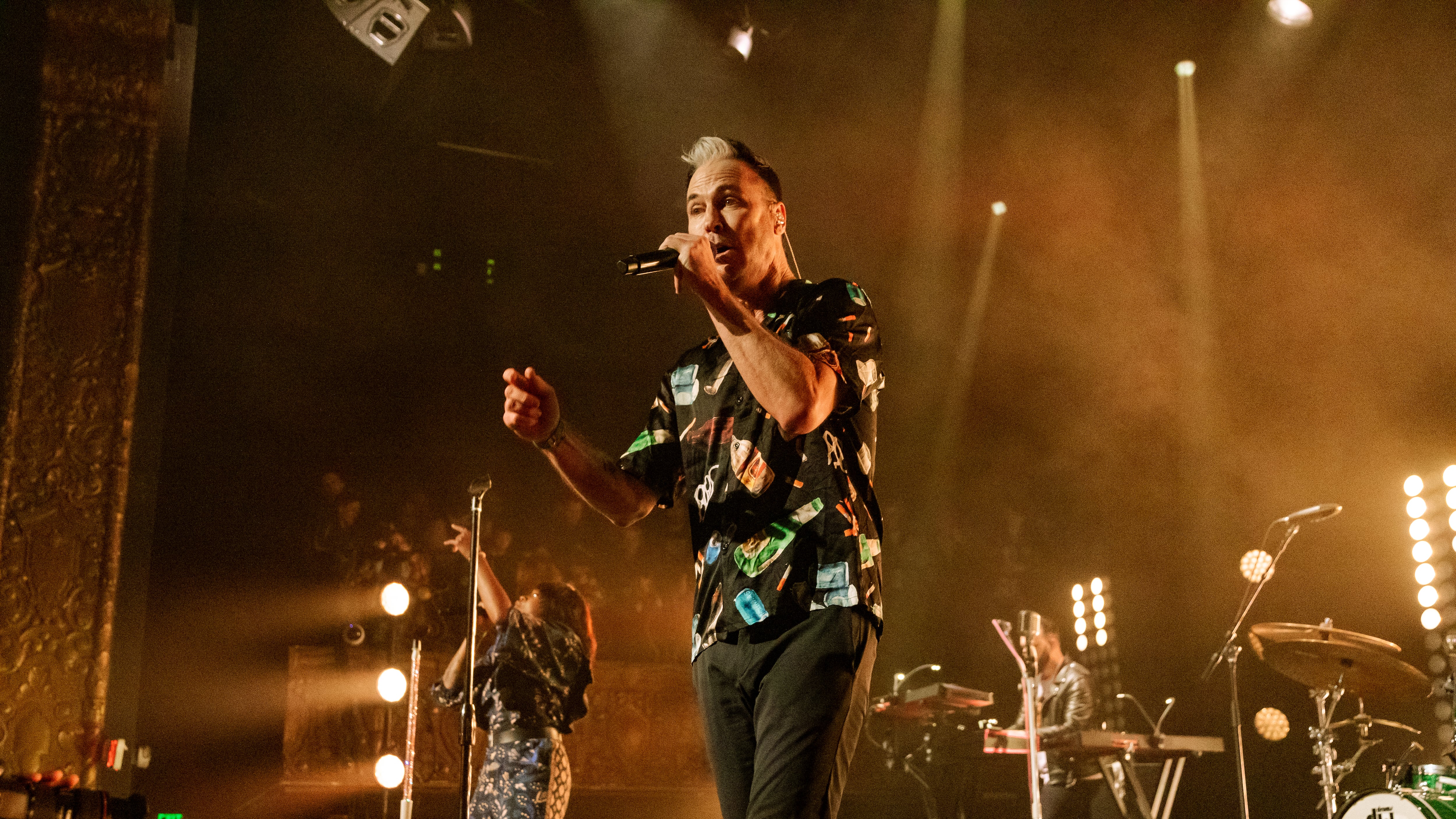 A photo of Fitzpatrick in the foreground singing into a microphone on stage. There are yellow lights in the background and the rest of the band can be seen in the background performing.