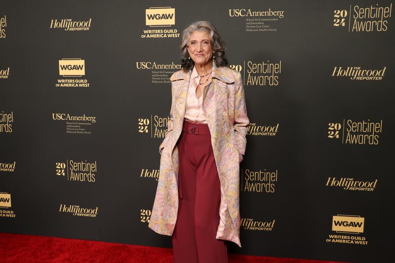 Amy has gray hair, a pink shirt and red pants on. She has a pink floral jacket on over.