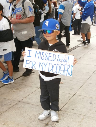 A young boy in a Dodgers hat smile while holding a sign that reads "I missed School For My Dodgers."