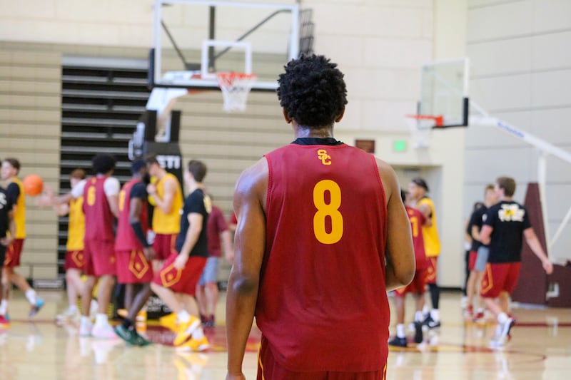 Jerry Easter II (8) stands with his back to the camera in the foreground wearing a cardinal USC basketball practice uniform with gold accents. The rest of the men's basketball team can be seen milling around under the basket in the background.