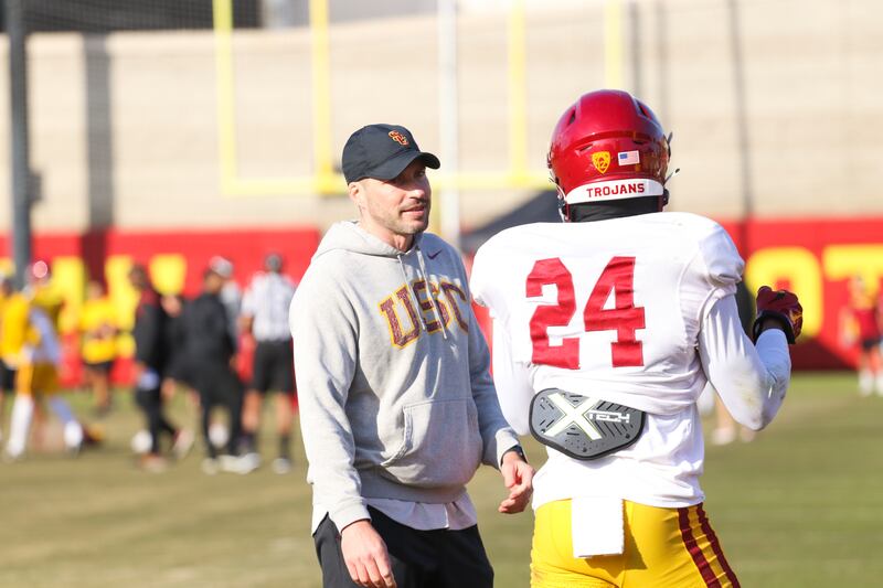 Alex Grinch is talking to a player in a cardinal helmet and white jersey with gold pants. Grinch is wearing a black hat with a gray sweatshirt.