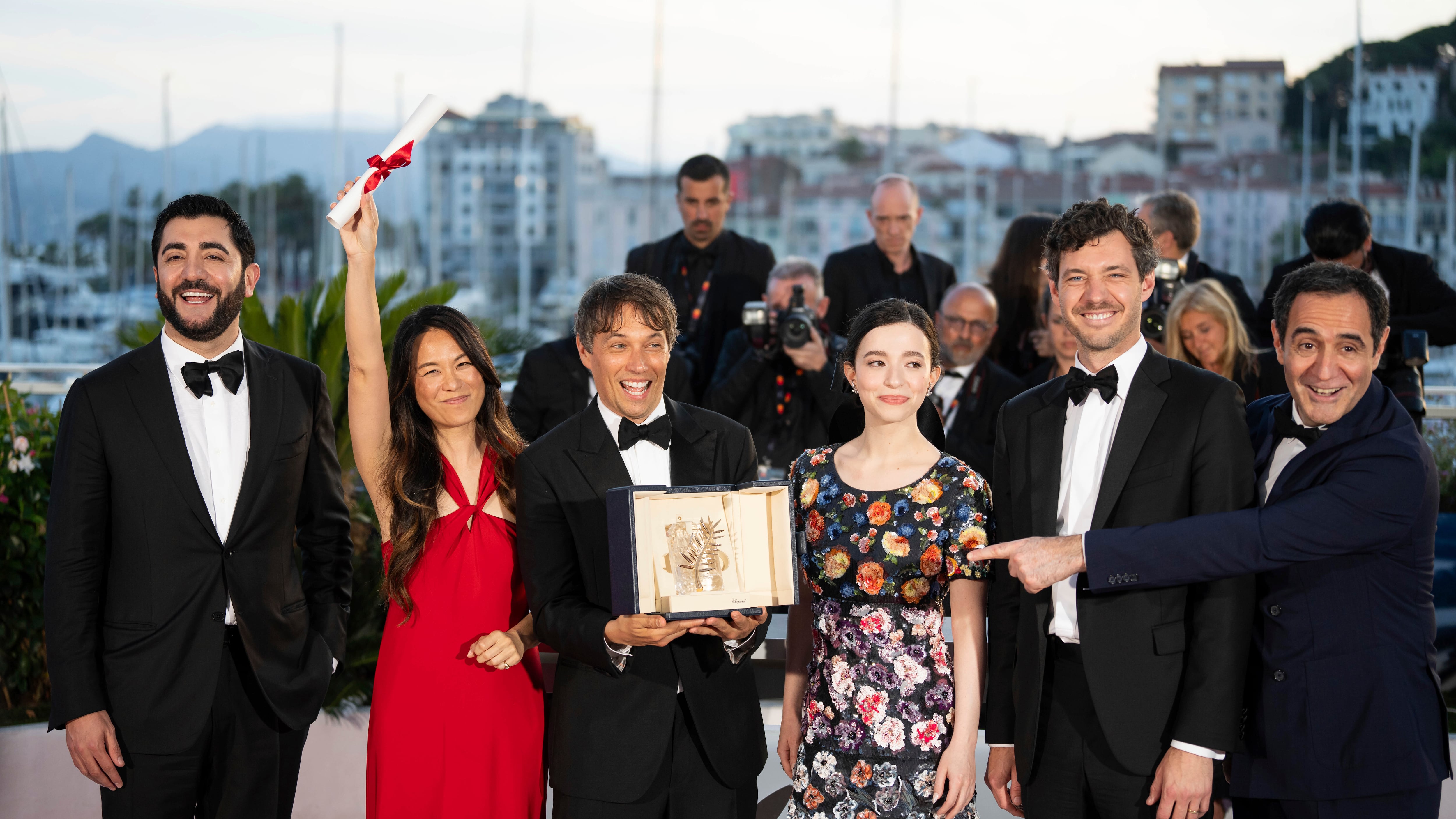 The men in the group are adorned in tuxedos. Mikey Madison wears a red dress and her arm is raised. Karren Karagulian wears a floral patterned dress. Sean Baker holds the Palme d'Or award in his hands with his mouth open, smiling.