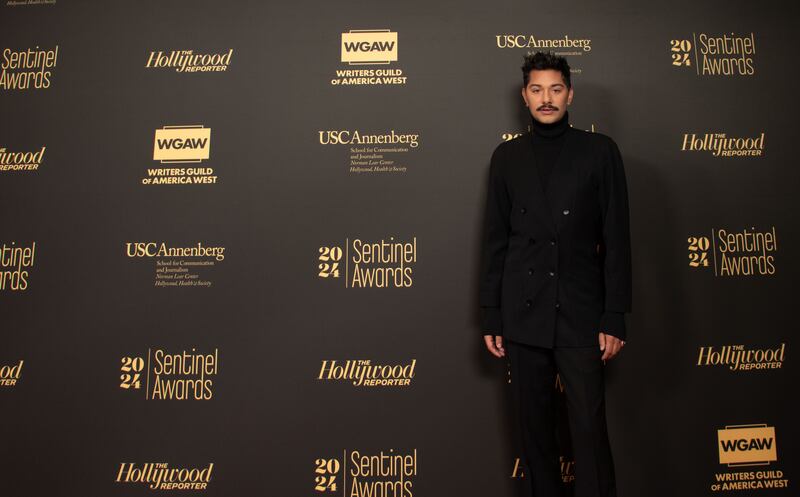 Mark is wearing a full length black suit, with a turtle neck on. He has a mustache and his hair is pointed upwards.