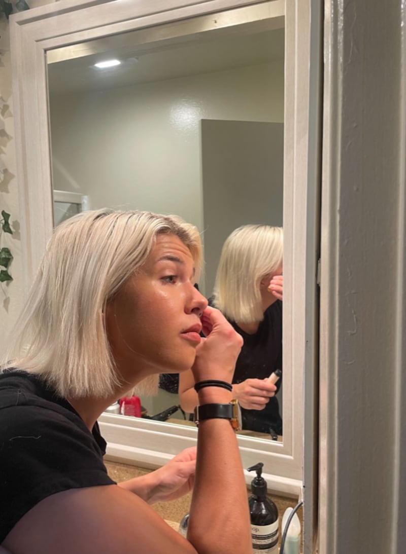 A women with icey blonde hair and a black shirt looked into a mirror while applying mascara