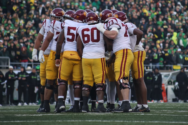 A USC football huddle at Autzen Stadium. The Trojans wear white jerseys and gold pants with cardinal accents and helmets. A crowd clad in black, green and yellow can be seen in the background.