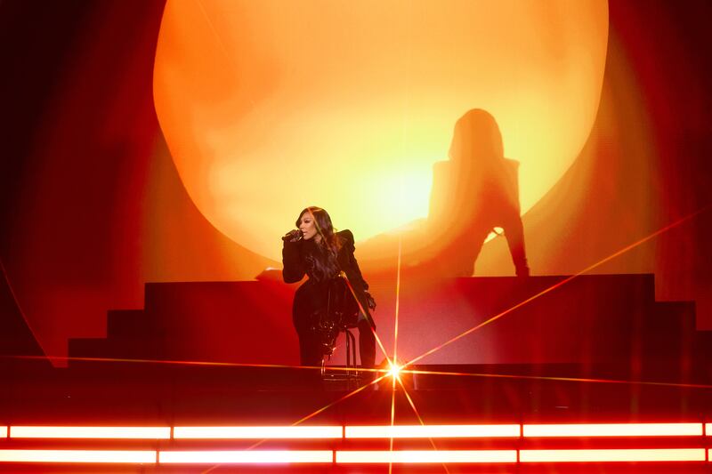 A woman is singing on stage by herself, with an orange glow in the background and a silhouette of a sitting woman behind her.