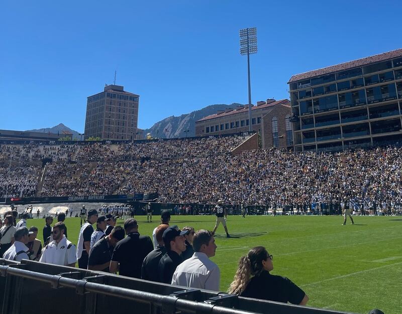 A field-side view, just steps from the pitch, of the entire Folsom Field stadium. The stands are packed for the game under a clear blue sky.