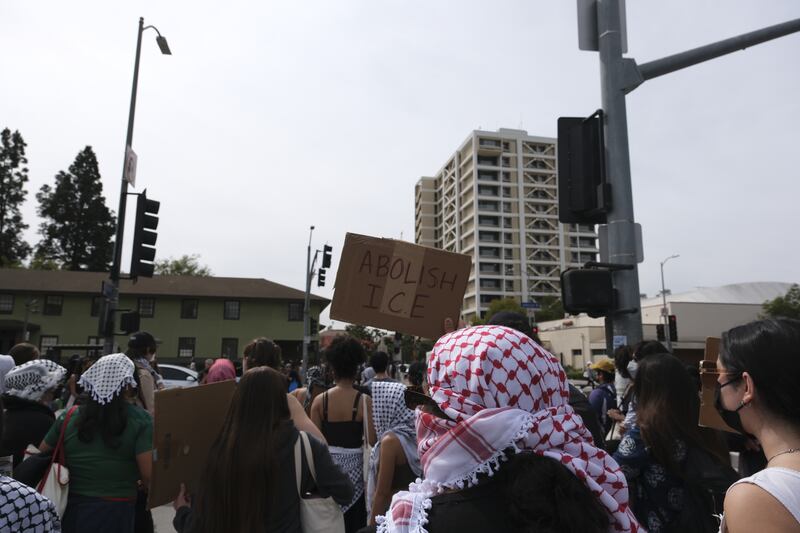 Protestors with masks on stand with their heads facing the camera. They are standing on a crosswalk to go into campus. On protestor holds a sign that reads "ABOLISH ICE."