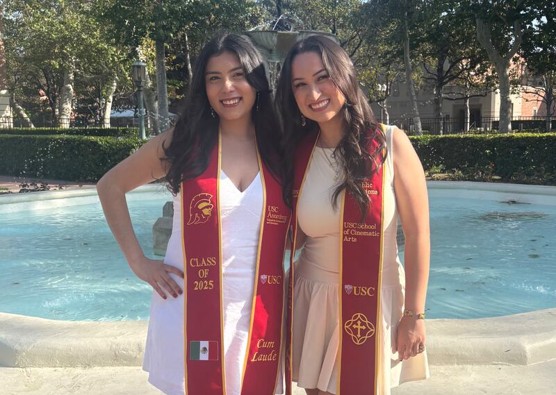 Two young women in white dresses with red sashes pose in front of a fountain.