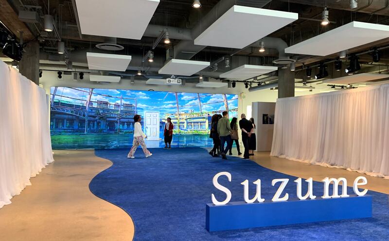 A photo of the exhibit of Makoto Shinkai’s past directorial work featuring a sign with the word "Suzume" and a blue carpet with white long drapes at the side of it.