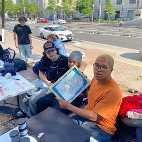 Photo of three people sitting at a table with one holding a screen printing device.
