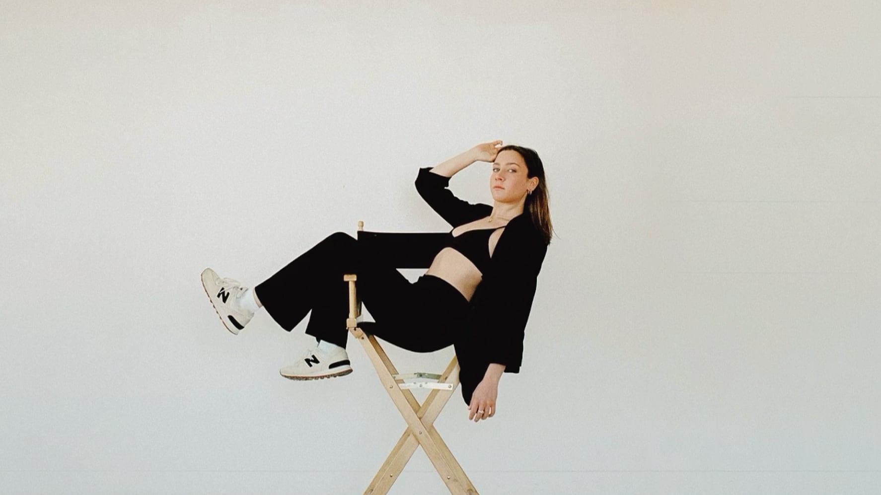 Against a solid white backdrop, Jamie lays across a directors chair. She is wearing white new balance sneakers, black pants bra and jacket. Her brown hair is down.