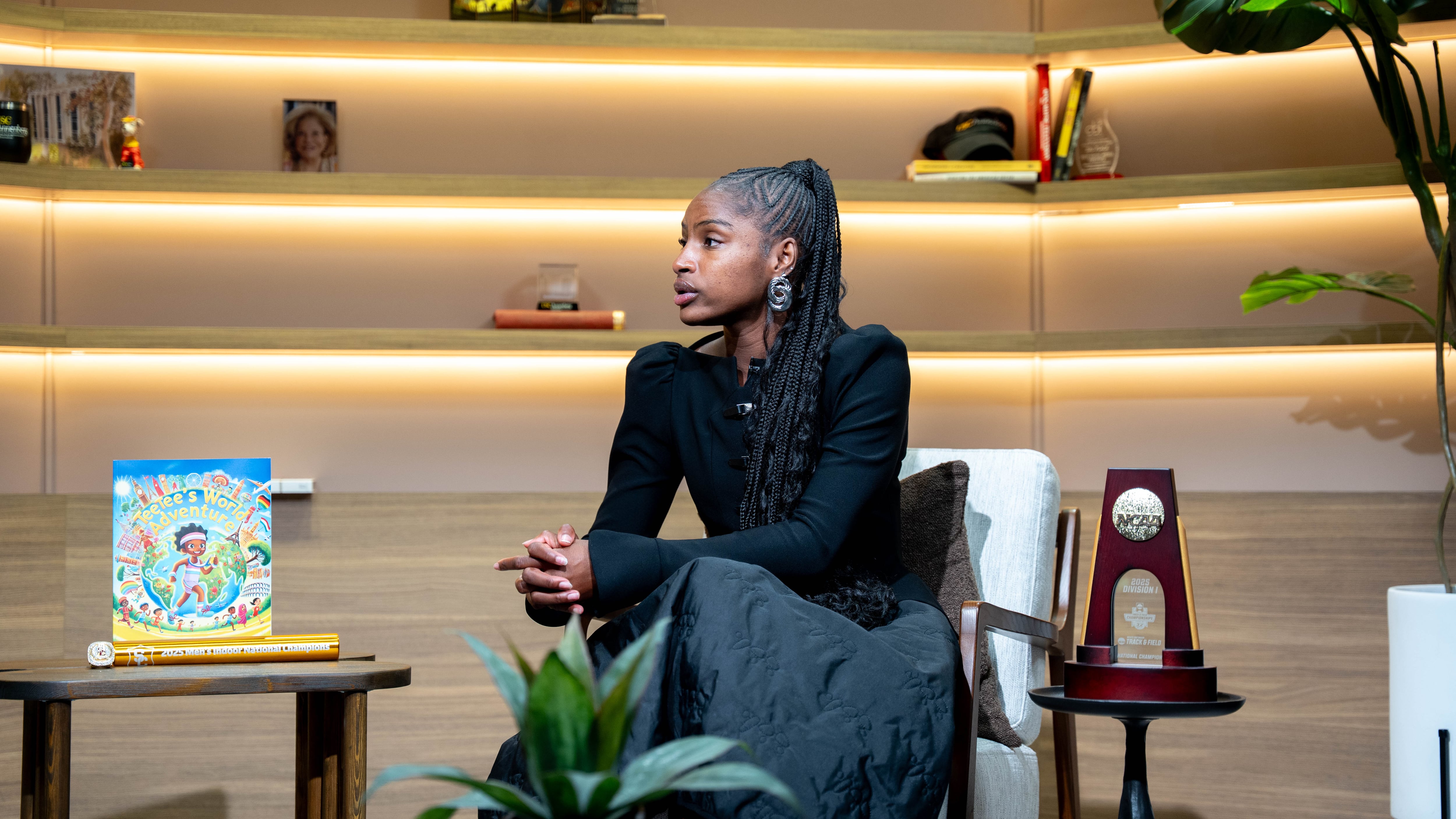 Black woman with braided hair sits in news room studio with a children's book on her left and NCAA trophy on her right.