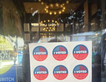 "I Voted" stickers in red, white and blue are displayed in a window; in the background are chandeliers with warm yellow bulbs hanging from a wood-beamed ceiling.