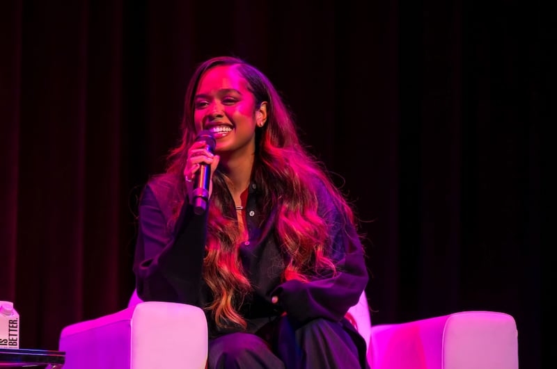 H.E.R. is holding a microphone while smiling. She is wearing and all black outfit and sitting in a white chair on stage.