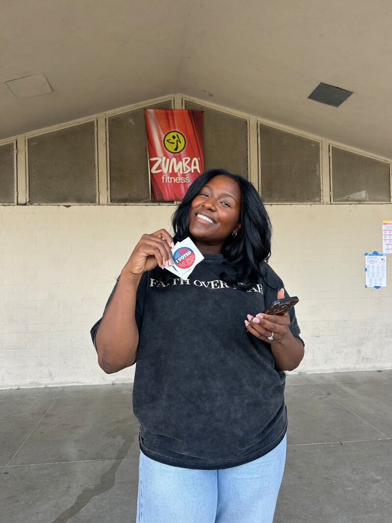 Photo of a woman in a grey shirt holding an "I voted" sticker.