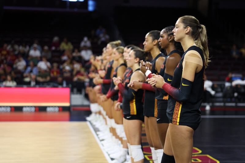 USC women's volleyball players stand at profile on court in a line. They wear black volleyball uniforms with cardinal and gold accents.
