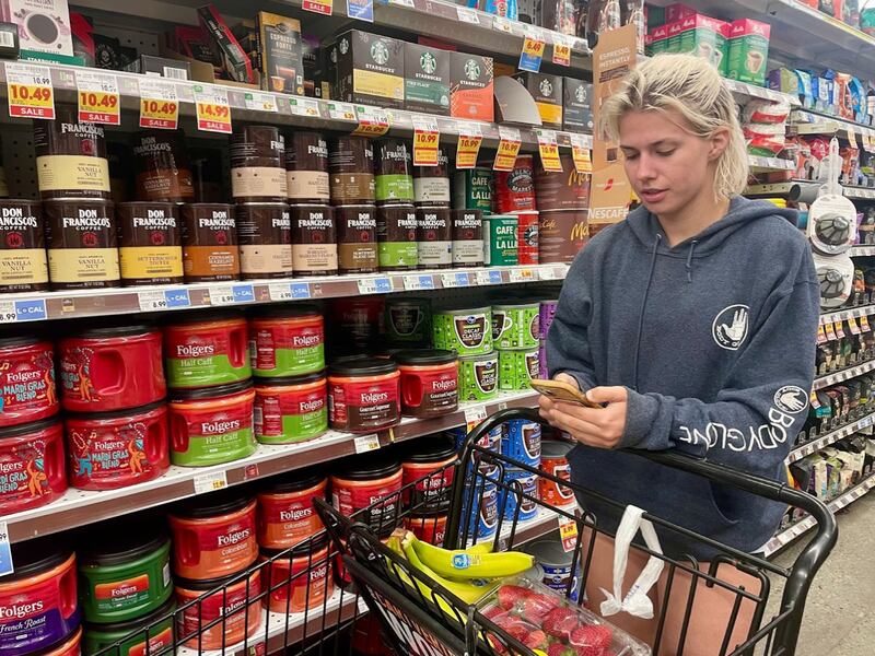 Wearing a blue sweatshirt, Light stands in a grocery aisle filled with coffee while checking her phone