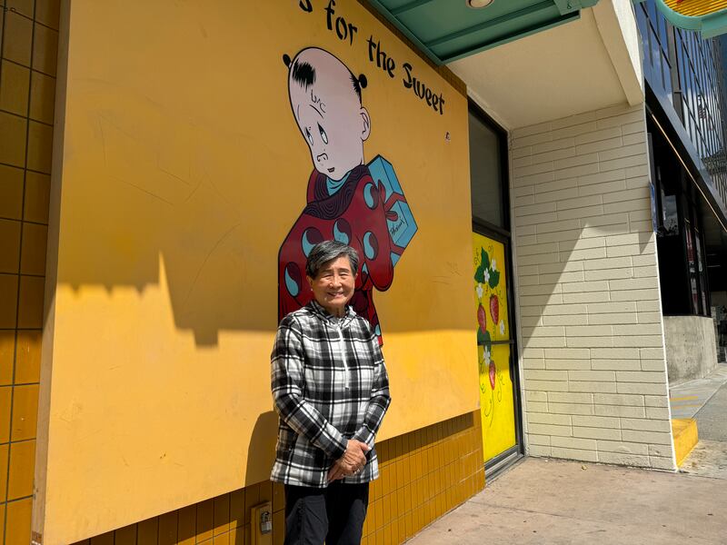 Photo of an older woman standing in front of a yellow wall with a painting of a child on it