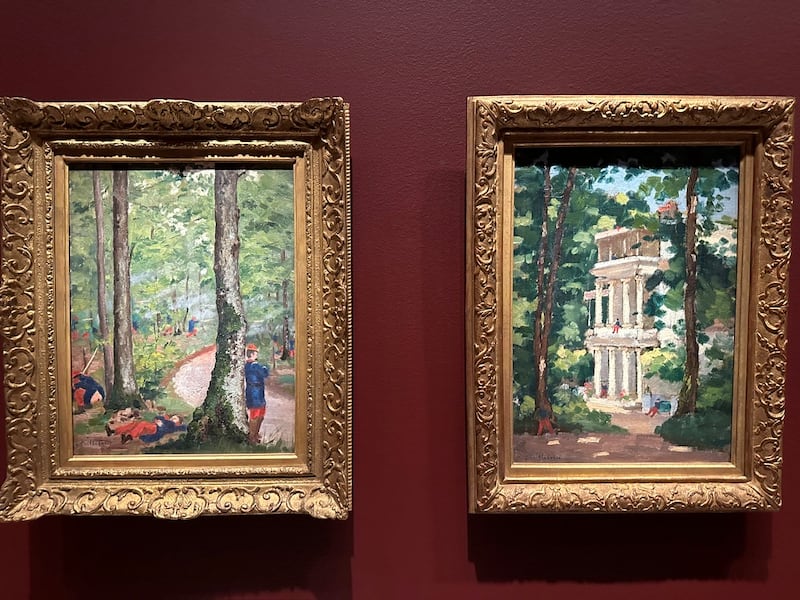 The two paintings are placed side-by-side. They are both in gold frames and against the dark red wall.