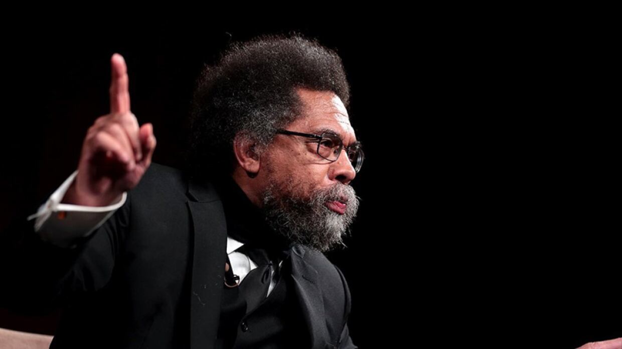 Cornel West in a black suit speaks with attendees and points his finger up