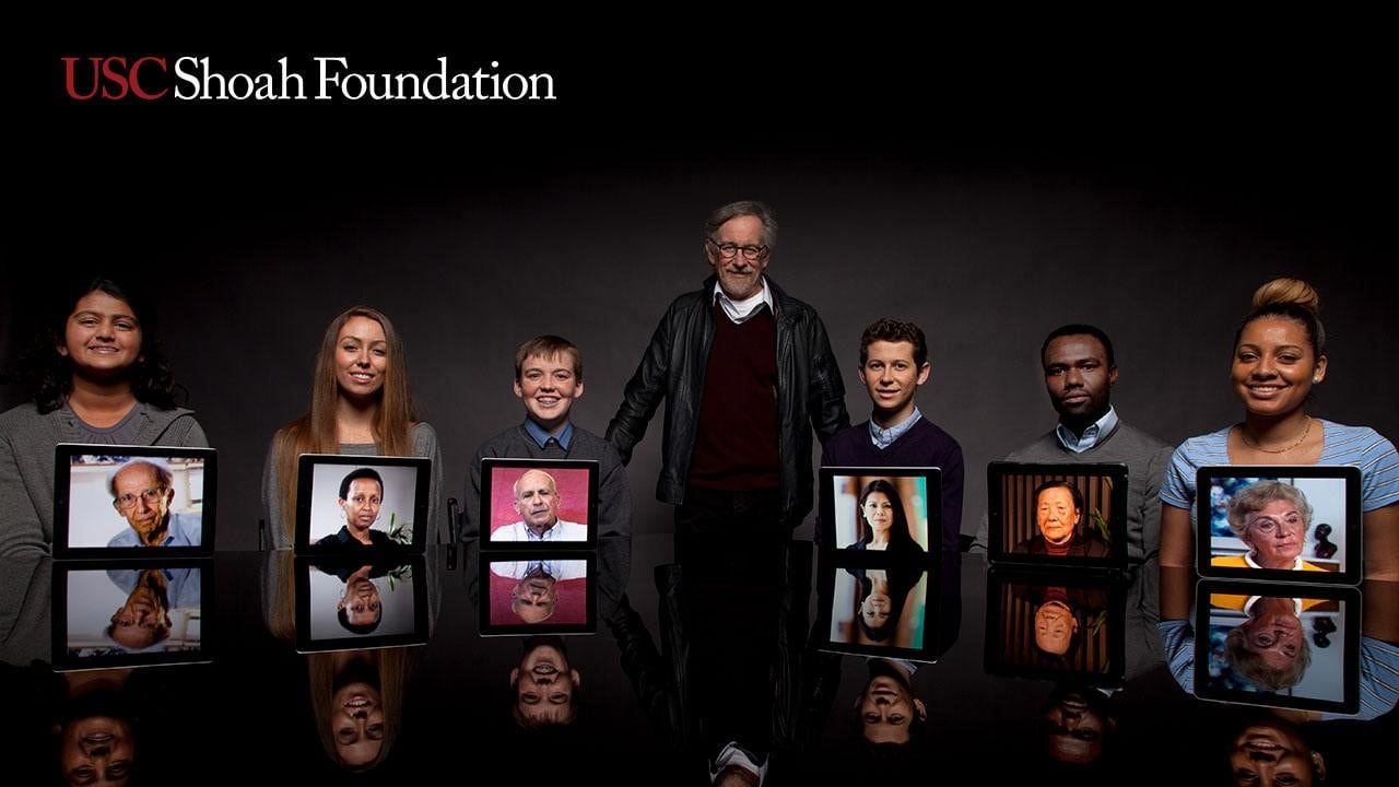 Steven Spielberg stands amongst a row of people with photos of USC Shoah Foundation testimonial participants.