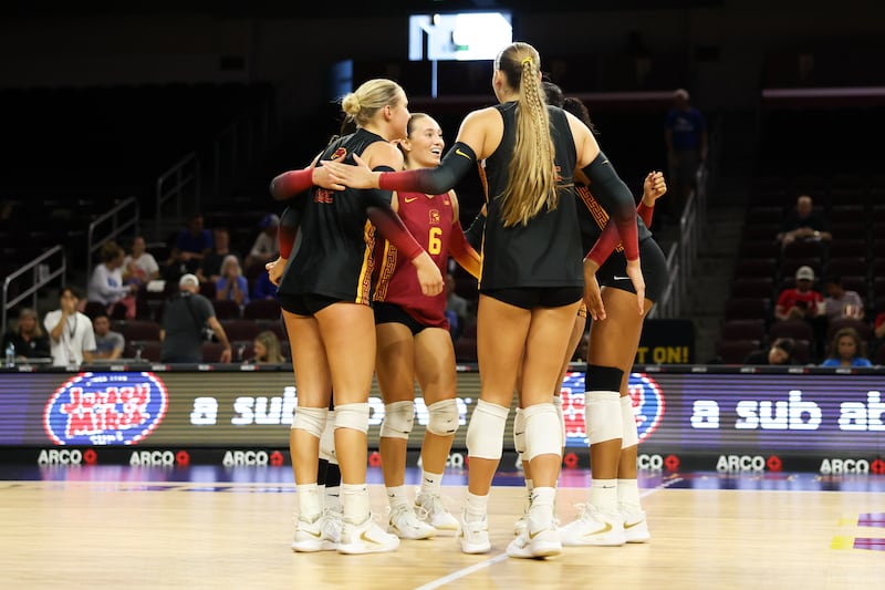 USC women's volleyball huddles on court, with a few smiling. They wear black and cardinal volleyball uniforms.