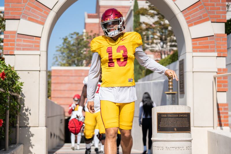 Williams (13) touches the Trojan Sword with his right hand as he emerges from the locker room before practice; he wears a gold USC football uniform with cardinal helmet.