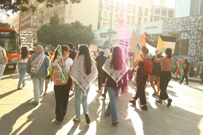 A group of people walking in the street, some holding protest signs and donning Keffiyehs.
