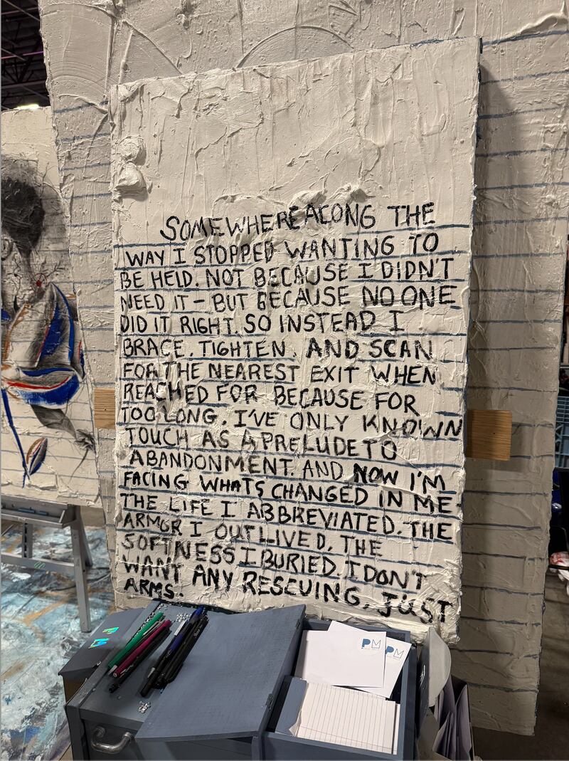 A textured white panel displays handwritten black text describing the loss of comfort with being held and the emotional armor formed through abandonment. Pens, cards, and paper sit on a small table below, inviting visitors to respond inside an indoor exhibition space.