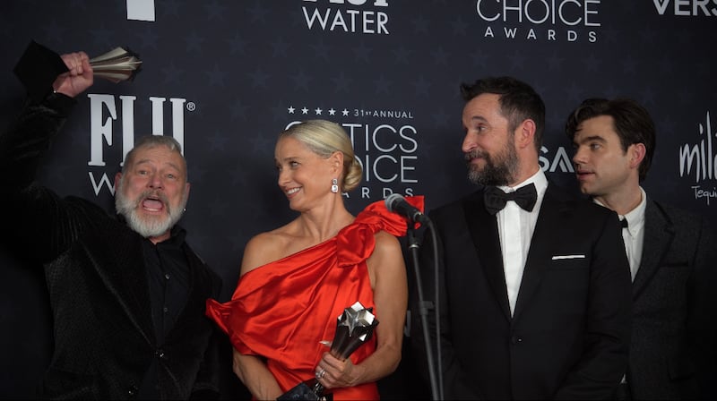A man on the left pumps up his trophy enthusiastically, while three other people look at him. The man is R. Scott Gemmill, the creator of "The Pitt" television series. Next to him is Katherine LaNasa who wears a red dress and is holding a trophy after winning Best Supporting Actress in a Drama Series. Next to her is Noah Wyle in a tuxedo, who won Best Actor in a Drama Series.