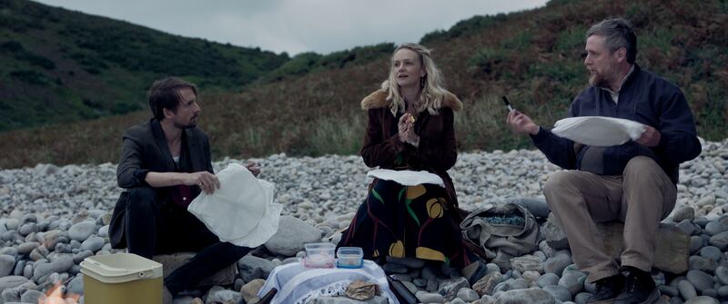 The three sit on white stones. There are gray mountains in the background. They look cold and are holding white sheets.