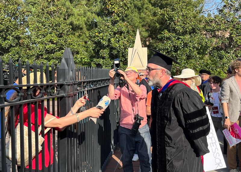 A professor in academic regalia speaks to press through a gate.