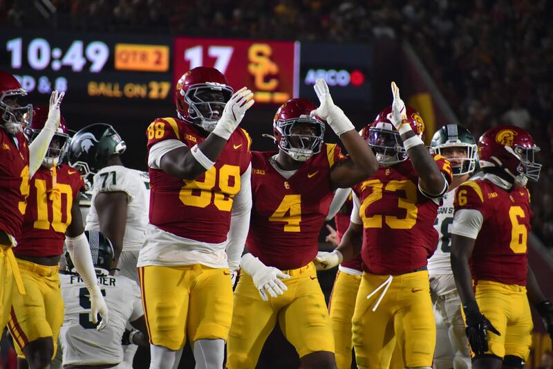 USC football players hold hands to their heads in imitations of shark fins; they wear cardinal and gold football uniforms.