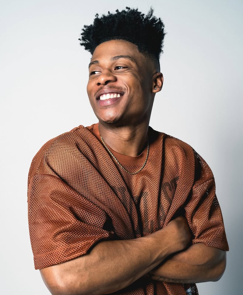 A studio portrait of Jared Smith, smiling, with short natural hair, wearing a brown textured shirt and a thin gold chain, arms crossed, against a light gray background.