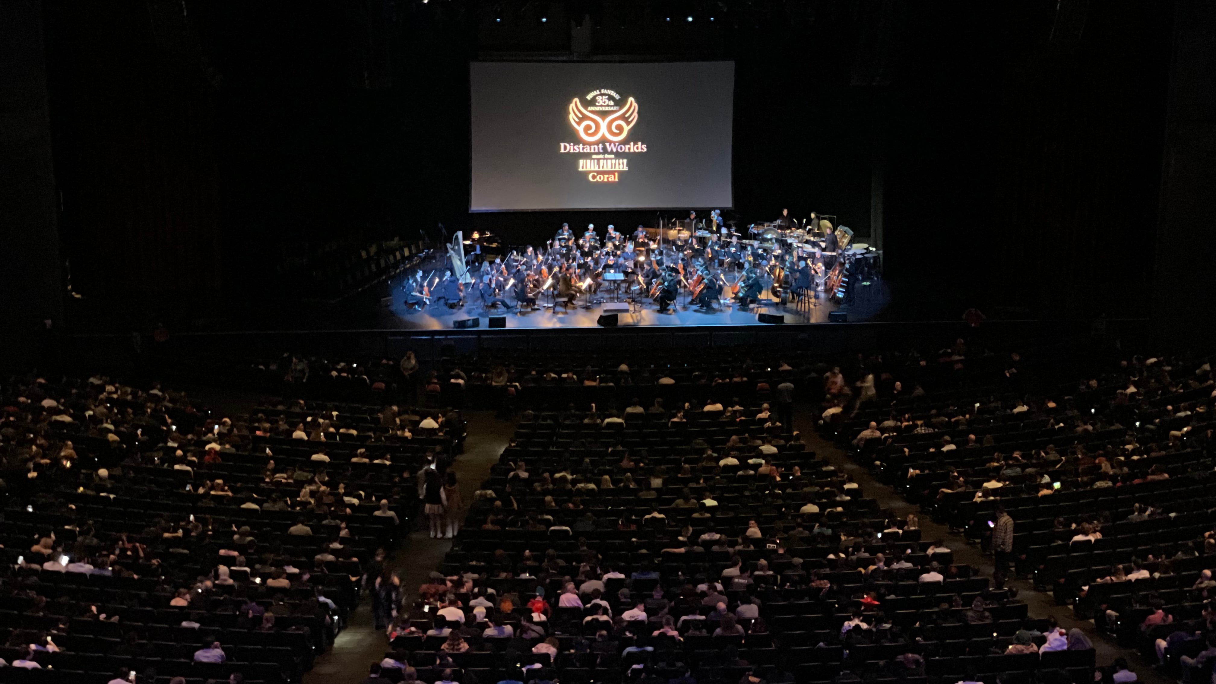 A photo of the audience and stage at the Microsoft Theater. The orchestra and choir are onstage and a projection on the stage reads: "Distant Worlds: Music from Final Fantasy Coral" with a logo.